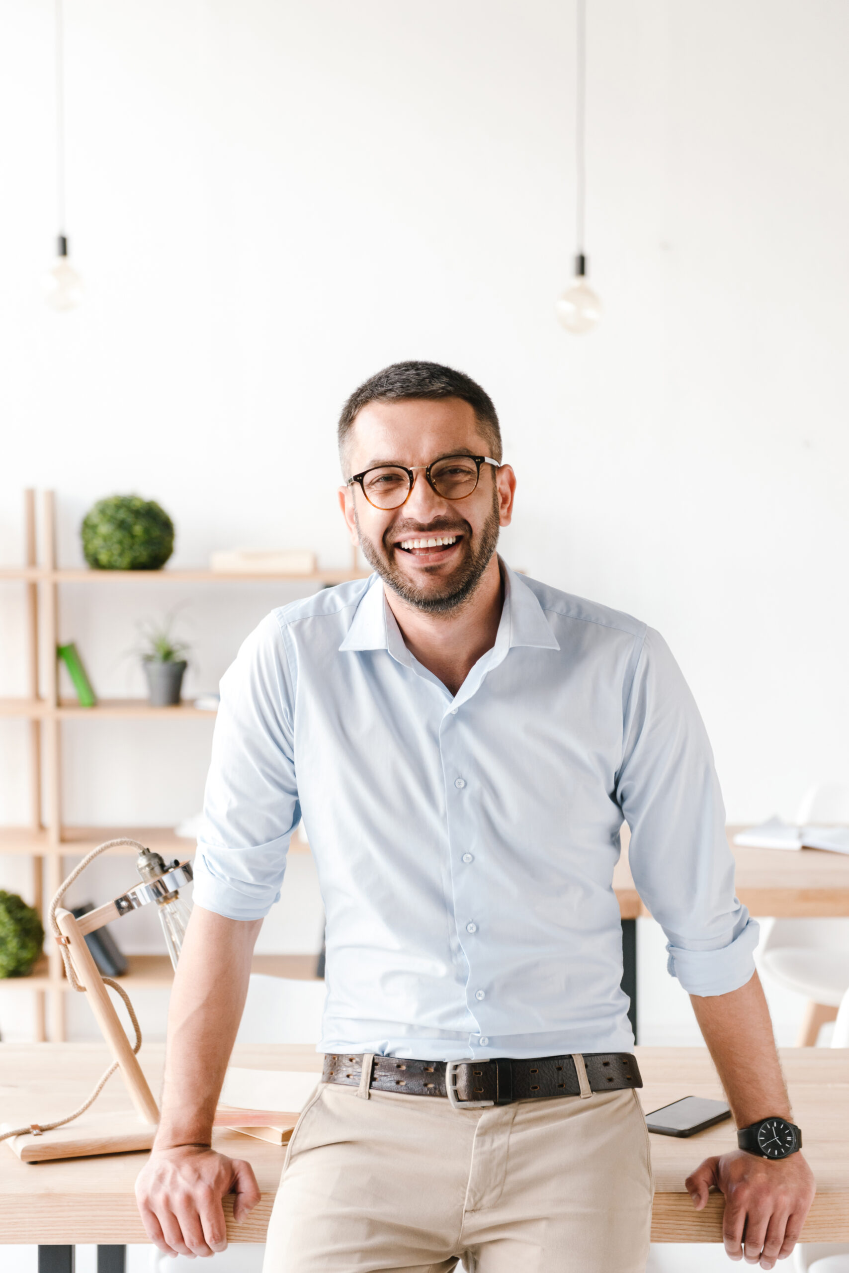 Happy portrait of businesslike man wearing white shirt sitting on table in office and smiling at camera