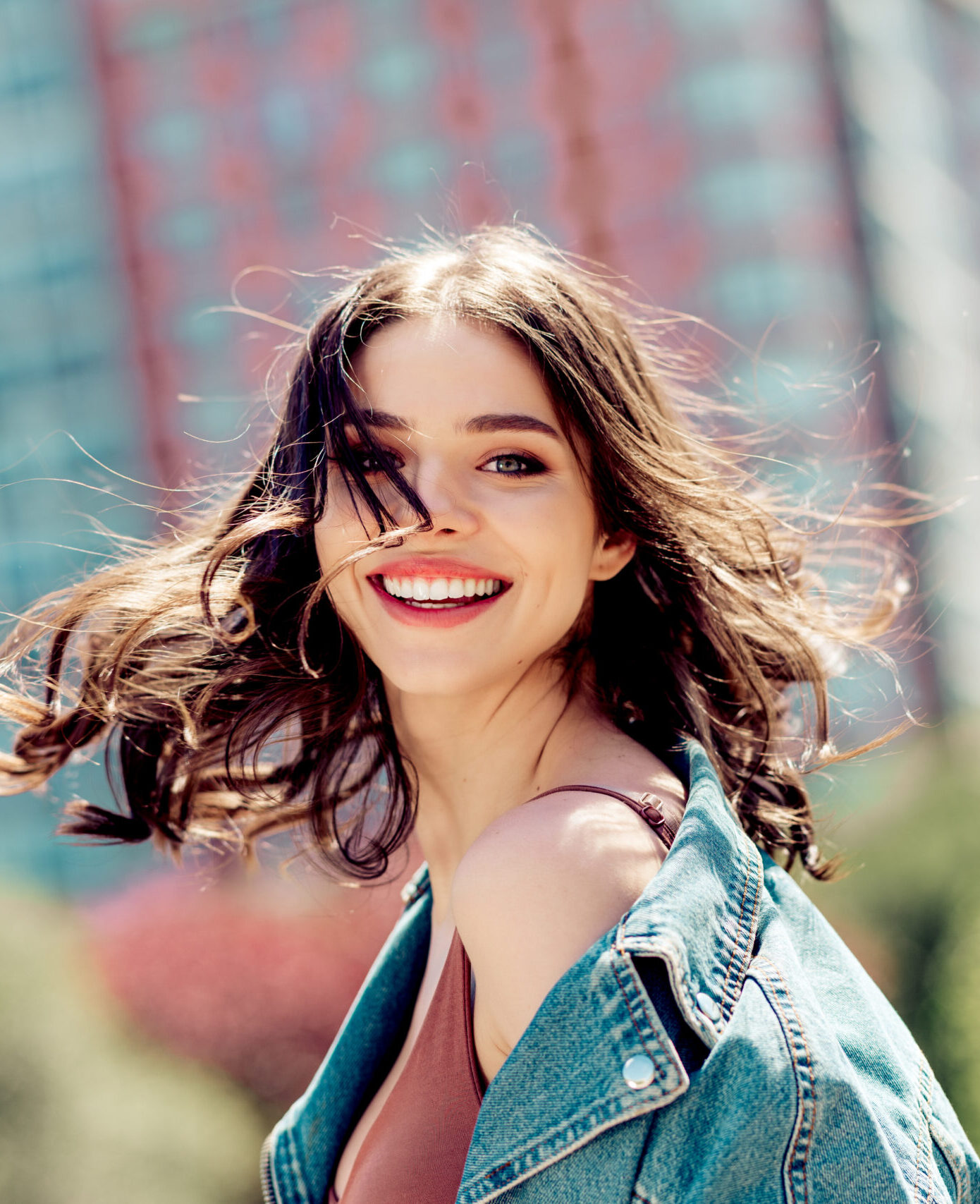 Smiling young woman in a denim jacket looks over her shoulder in a candid outdoor city portrait. Windblown hair, bright smile and sunlit skin convey joy, freedom and youthful energy.